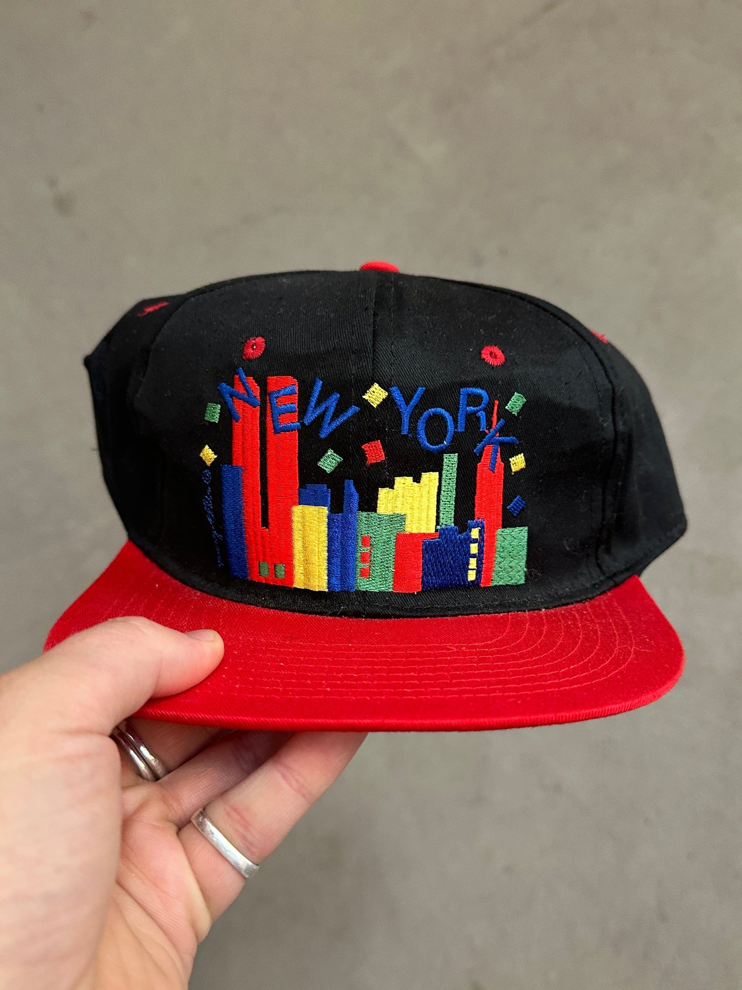 Black and red cap with colorful 'New York' design held by a hand against a gray background