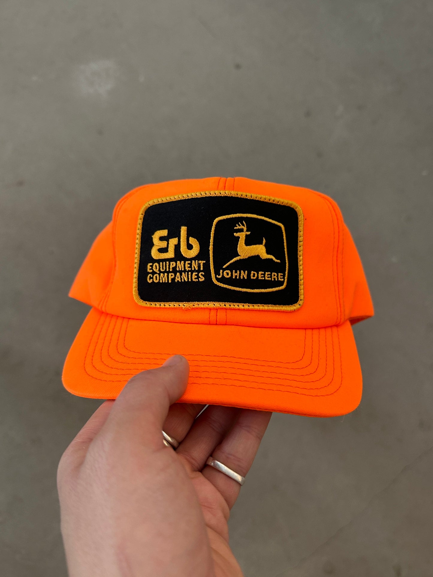 Orange cap with John Deere logo held by a hand against a gray background