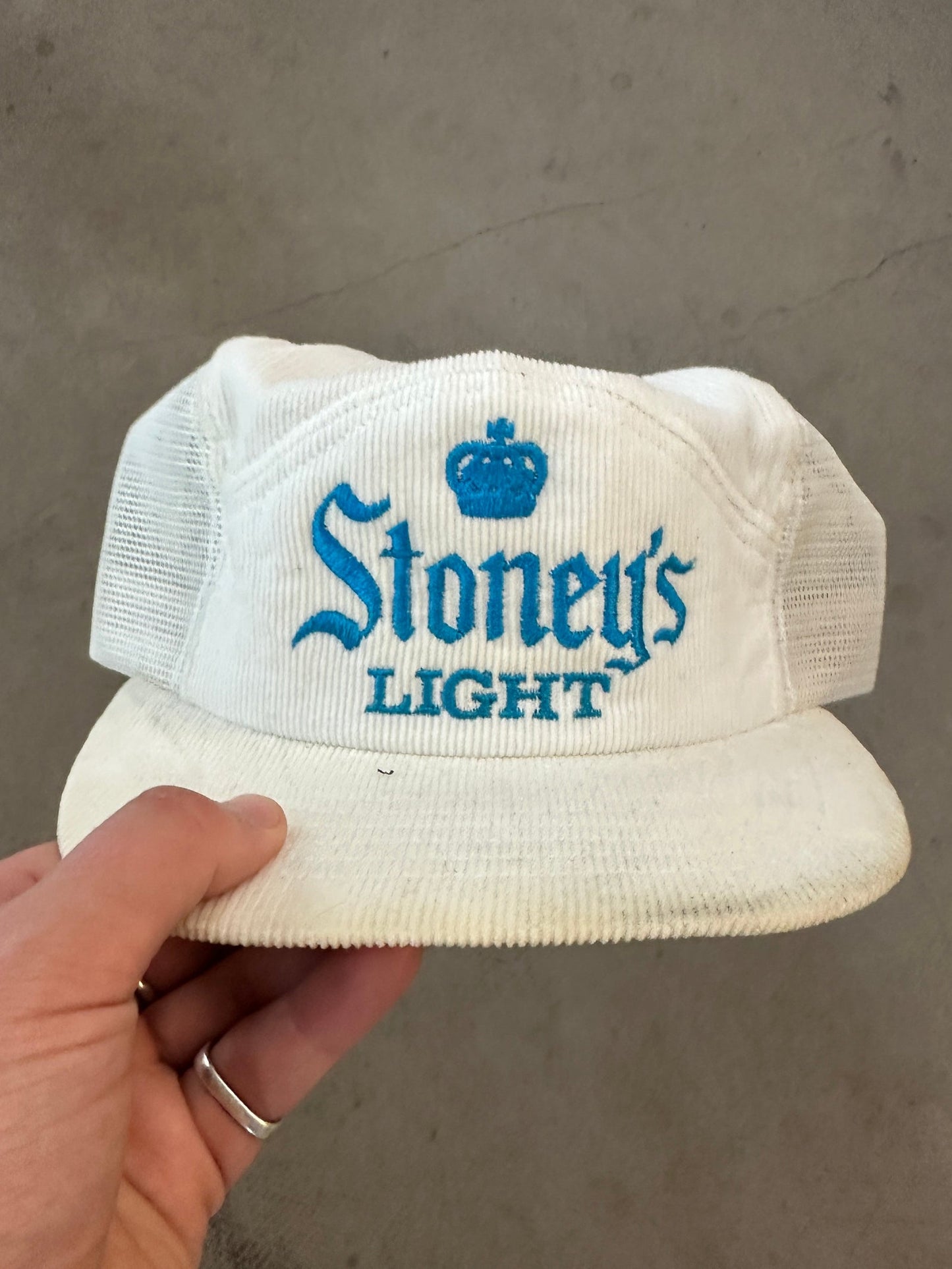 White cap with 'Stoney's Light' branding held by a hand against a gray background