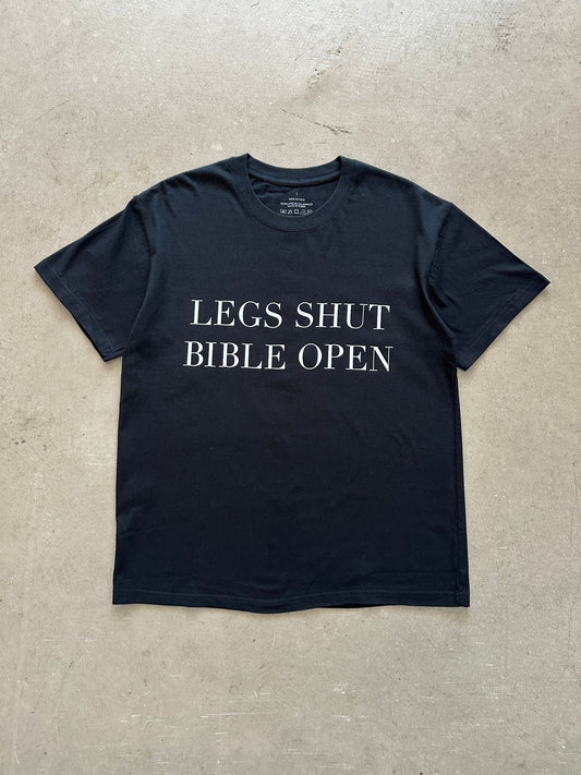 Black t-shirt with text 'LEGS SHUT BIBLE OPEN' on a concrete surface