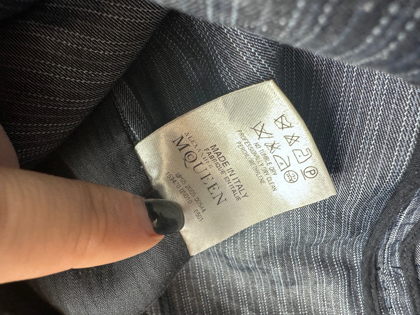 Close-up of a clothing label reading McQueen, attached to a gray striped fabric garment, with a finger pointing at it.