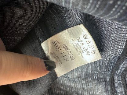 Close-up of a clothing label reading McQueen, attached to a gray striped fabric garment, with a finger pointing at it.