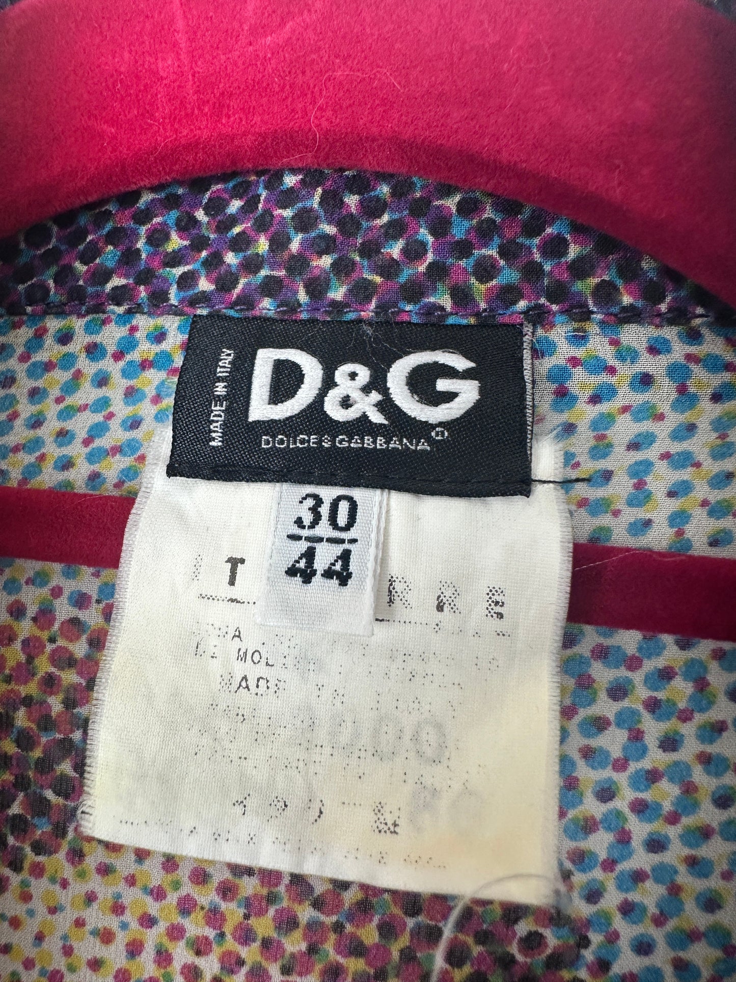 Close-up of a Dolce & Gabbana garment label showing the D&G logo on a black tag next to a size 30/44 paper label, with a pink fabric edge and a multicolored patterned textile background.