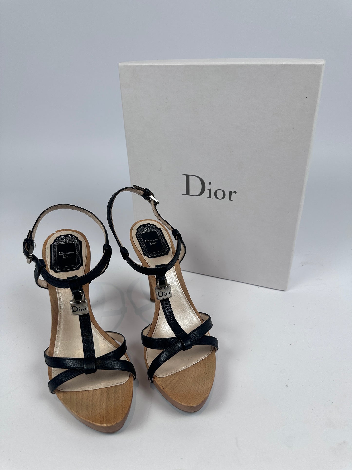A pair of Dior strappy high-heeled sandals with black leather straps and beige wood-look soles, displayed next to a white Dior shoebox on a light gray background.