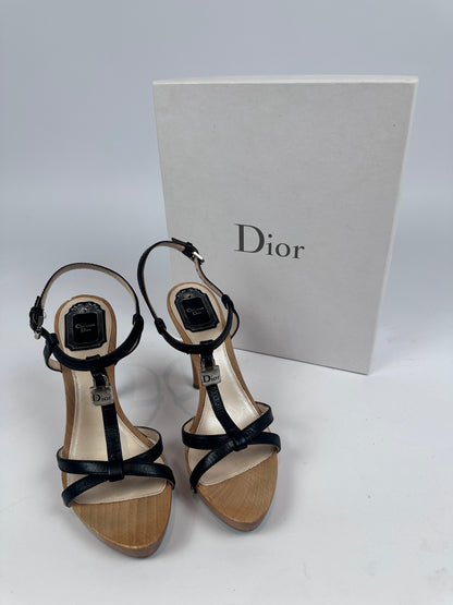A pair of Dior strappy high-heeled sandals with black leather straps and beige wood-look soles, displayed next to a white Dior shoebox on a light gray background.