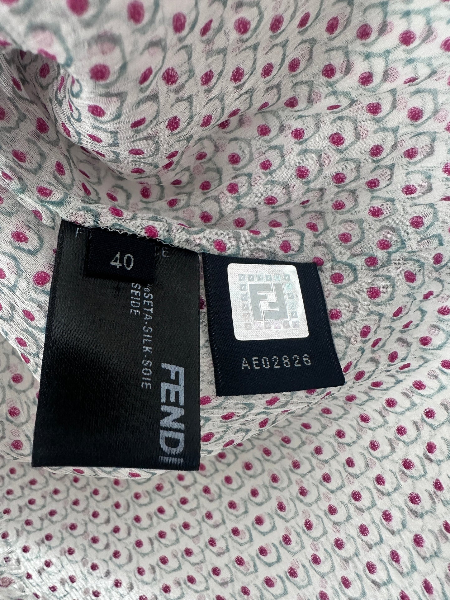 Close-up of a Fendi clothing tag sewn onto patterned fabric. The tag is black with white text reading FENDI and size 40, while the fabric has a white base with pink and gray floral motifs.