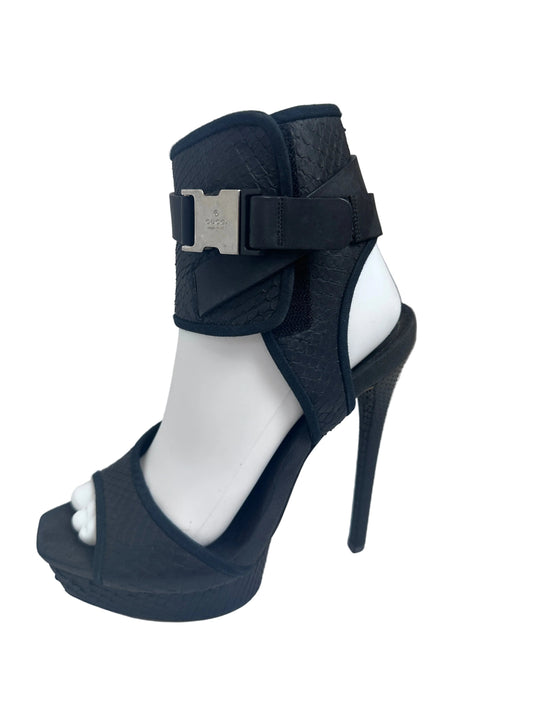 A black high-heeled platform sandal with an ankle strap and silver buckle, displayed on a white mannequin leg.