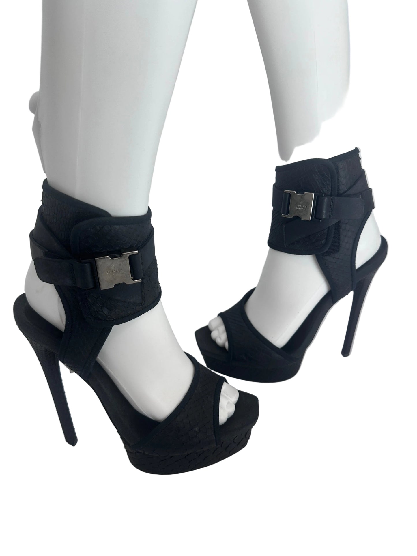 A pair of black high-heeled sandals displayed on white mannequin legs against a plain light background.
