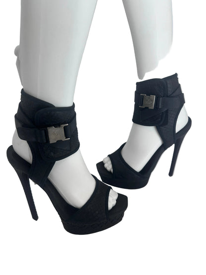 A pair of black high-heeled sandals displayed on white mannequin legs against a plain light background.