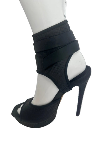 A black high-heeled platform sandal with wraparound ankle straps, displayed on a white mannequin leg against a light gray background.