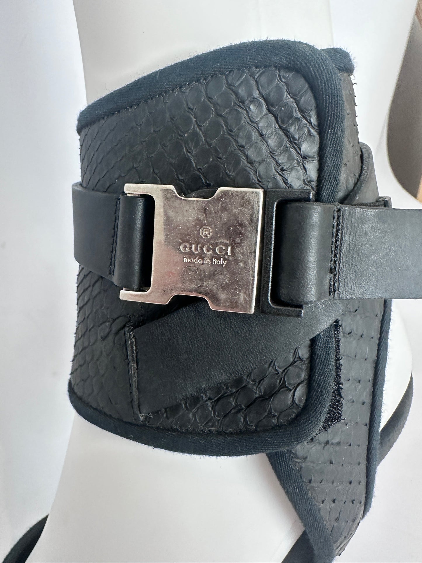 A black Gucci leather ankle cuff featuring a crocodile-embossed texture and a prominent silver-tone metal buckle engraved with 'GUCCI Milano & Italy'.