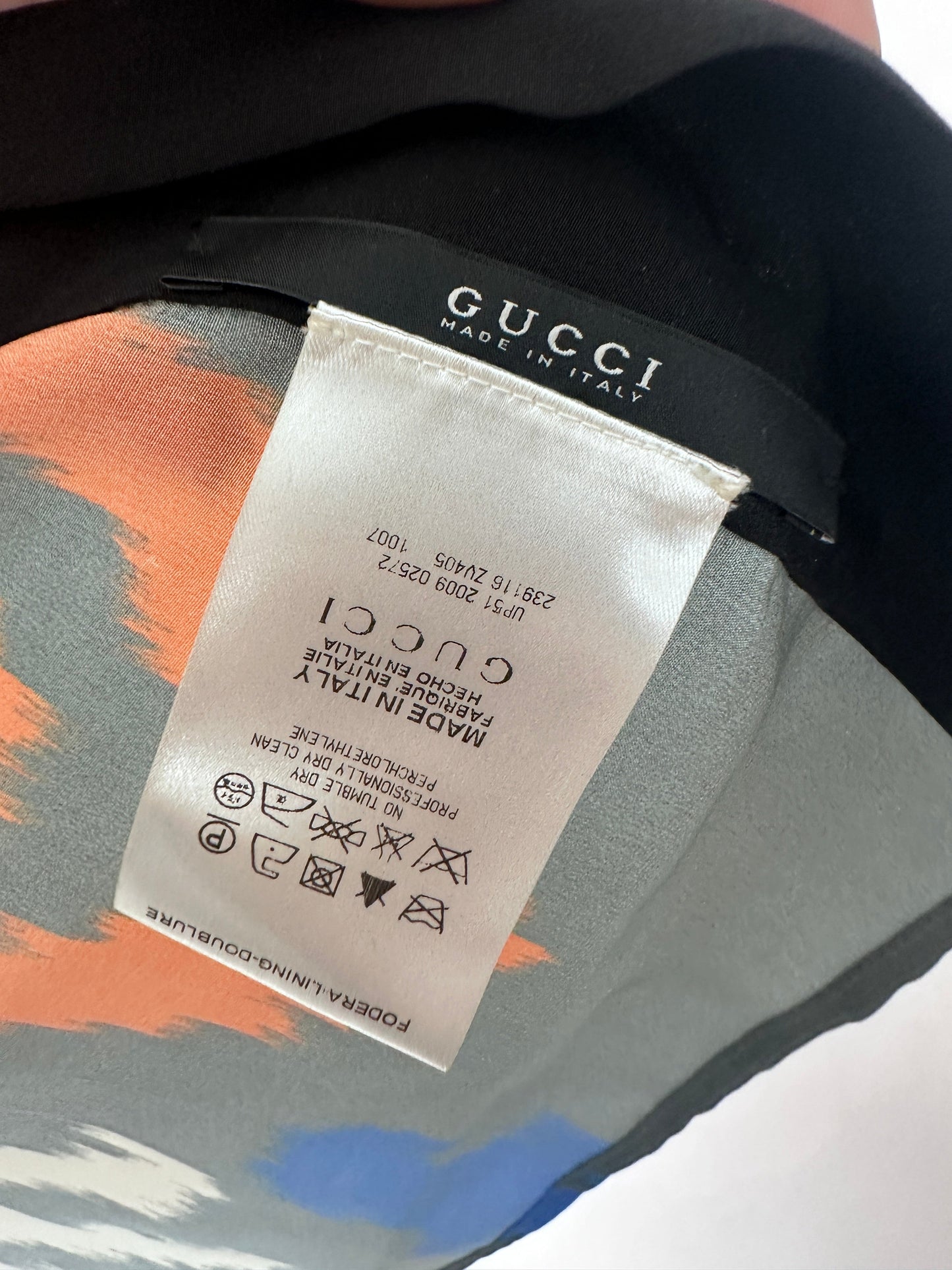 Close-up of a Gucci garment label sewn onto a black Gucci strap, with part of the patterned fabric visible underneath in gray, orange, and blue.