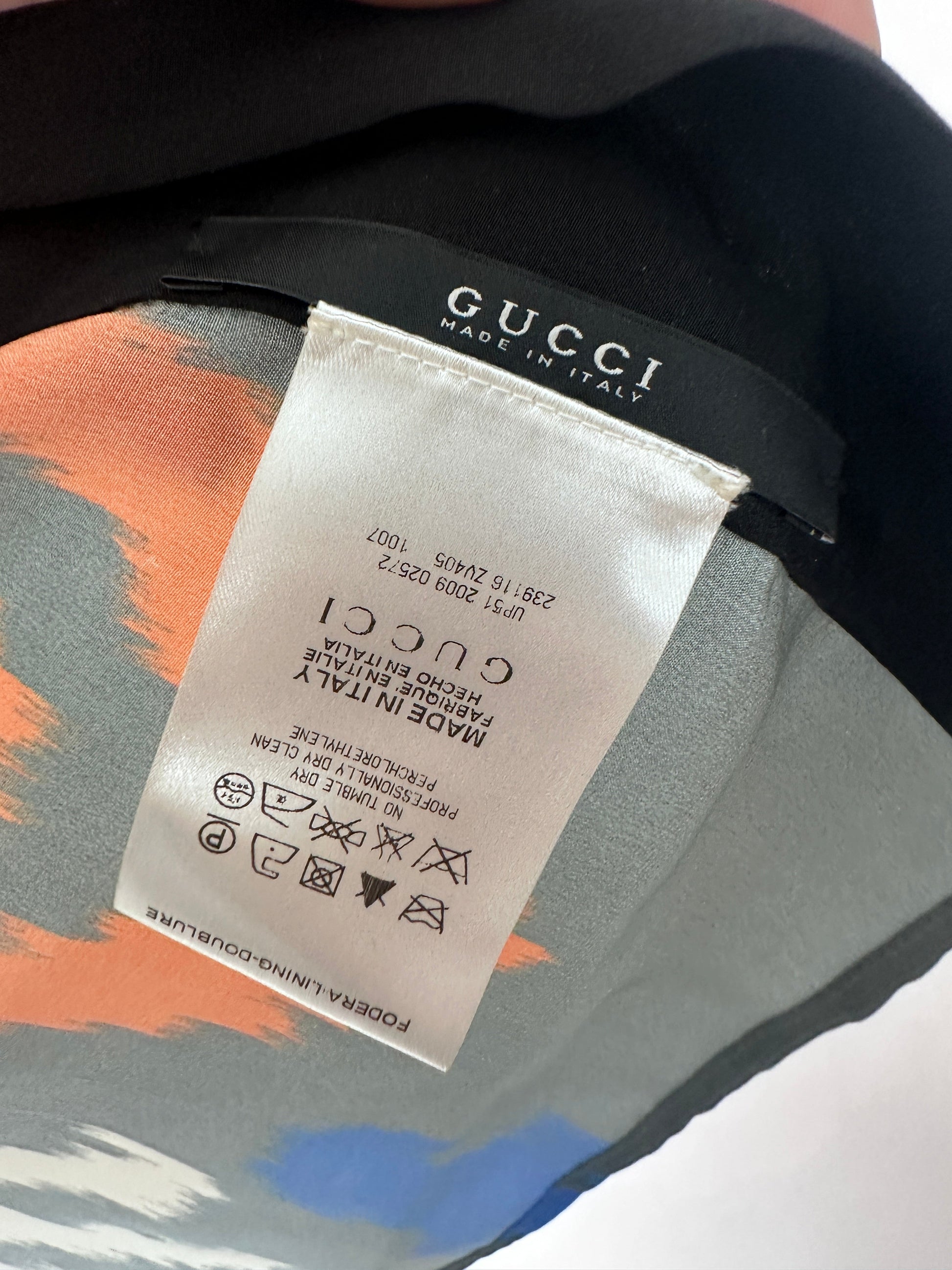 Close-up of a Gucci garment label sewn onto a black Gucci strap, with part of the patterned fabric visible underneath in gray, orange, and blue.