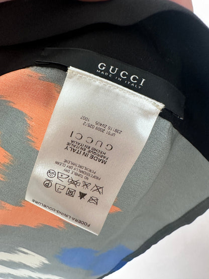 Close-up of a Gucci garment label sewn onto a black Gucci strap, with part of the patterned fabric visible underneath in gray, orange, and blue.