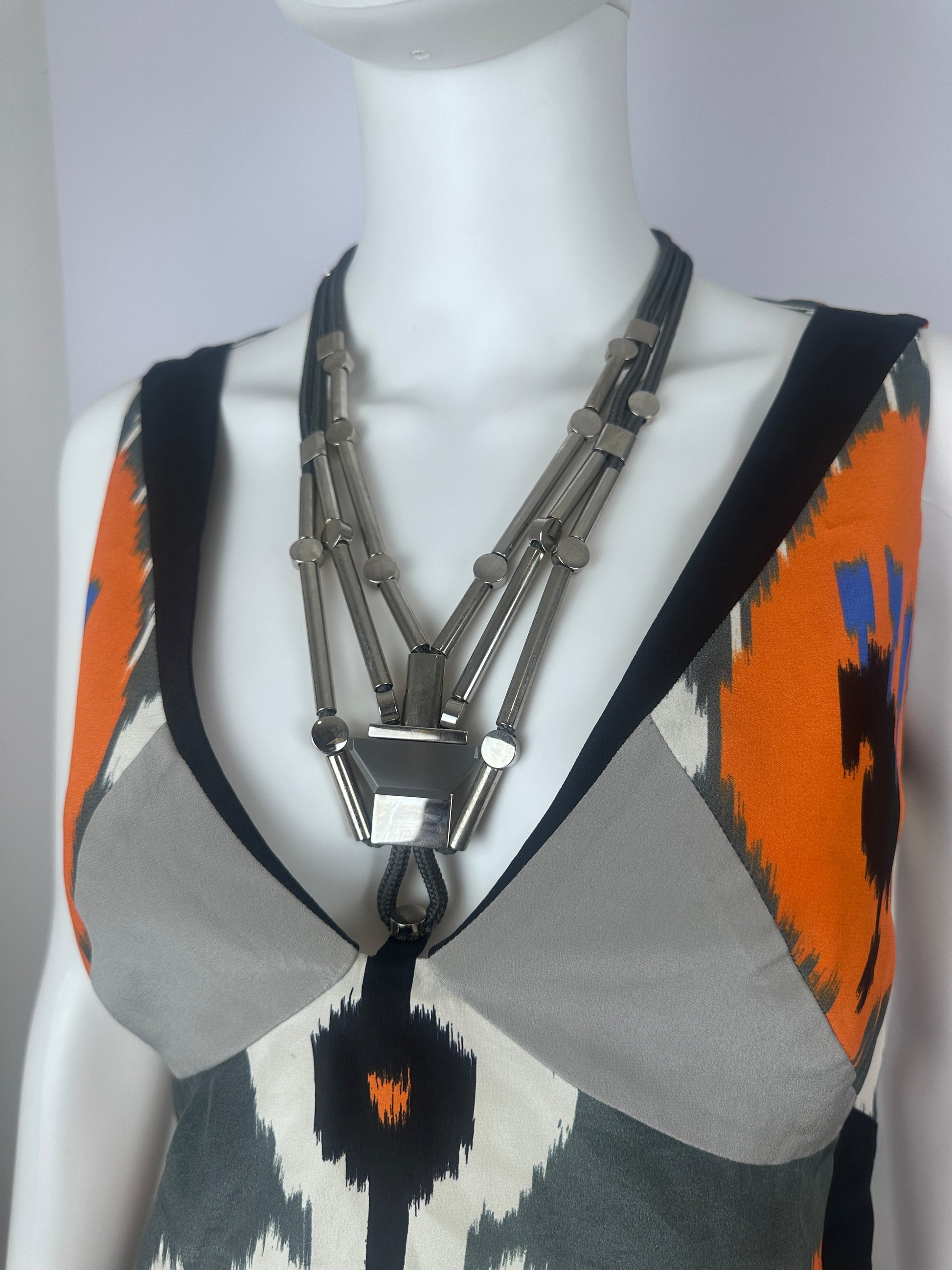 A close-up of a garment displayed on a white mannequin torso, featuring a bold geometric pattern with orange, black, gray, and white sections, accented by a prominent silver-toned metal chain detail across the front.