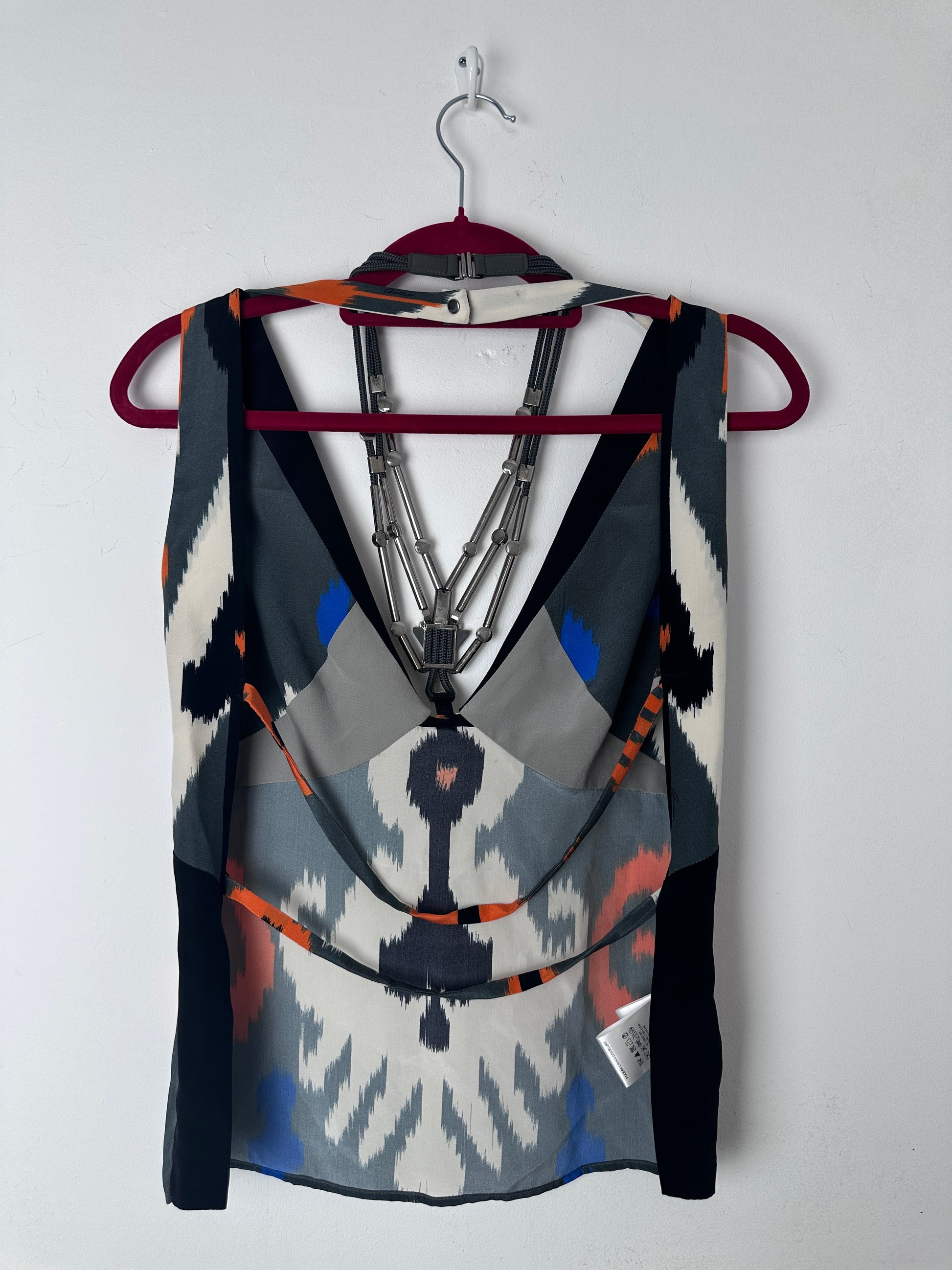 A folded patterned scarf or shawl draped over a hanger against a plain light background. The fabric features a geometric ikat-inspired design in black, white, gray, blue, and orange, with a small white brand tag visible on the right side.
