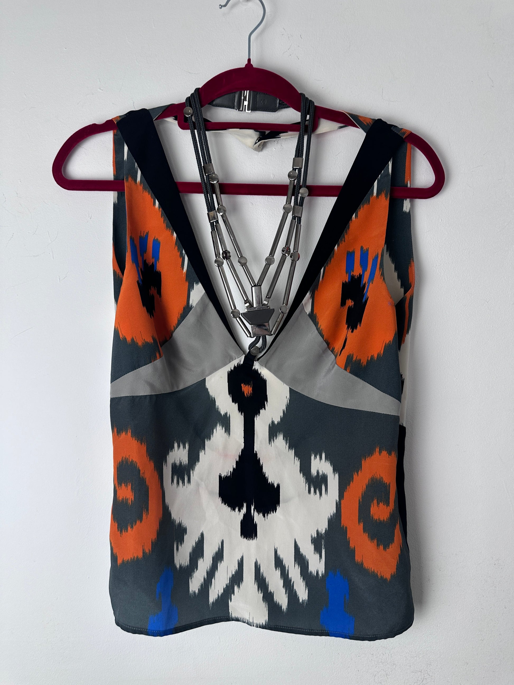 A patterned fabric pouch or bag with a geometric ikat design in gray, orange, white, black, and blue, featuring a silver metal zipper and a dark red strap, shown on a white background.