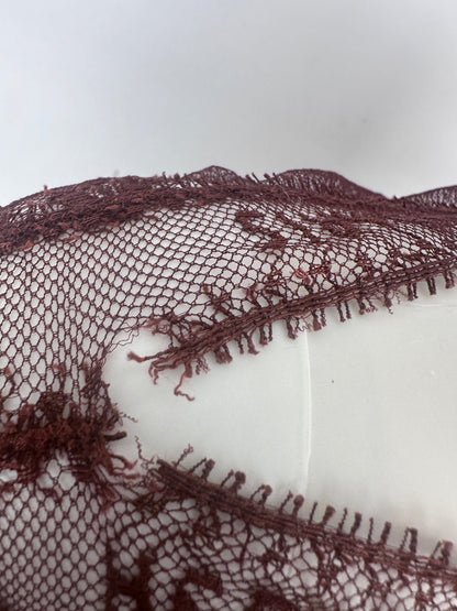 A close-up of a dark plum lace fabric with a V-shaped cutout and frayed, tassel-like trim along the edges, displayed on a white mannequin torso against a light gray background.
