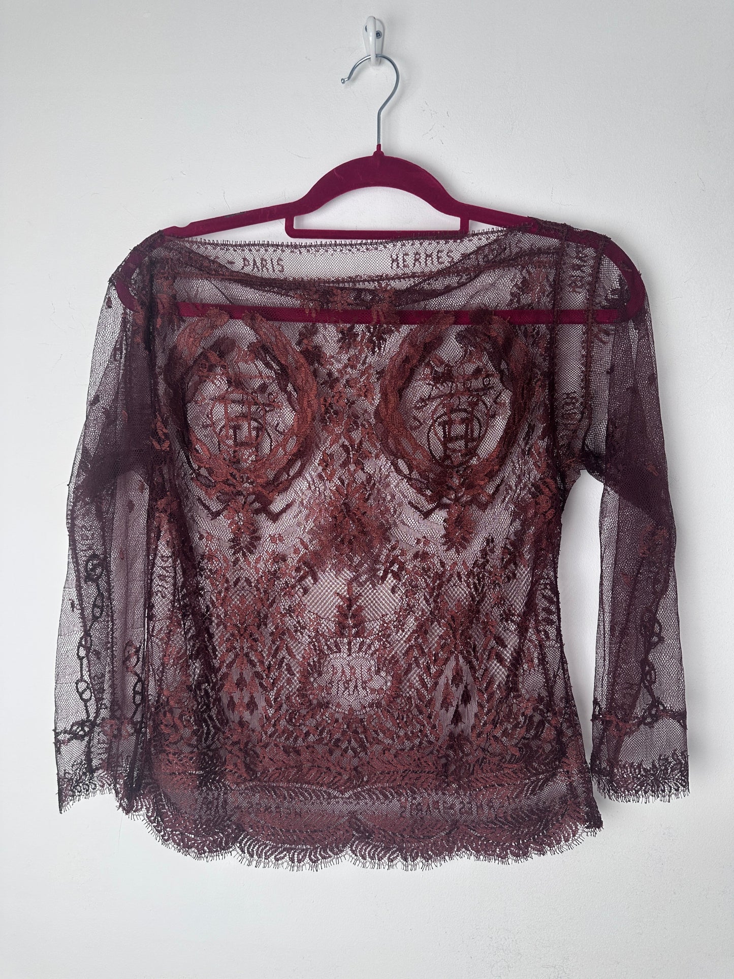 A sheer burgundy lace shawl with a floral pattern and fringed edges, draped over a matching burgundy hanger against a plain white background.