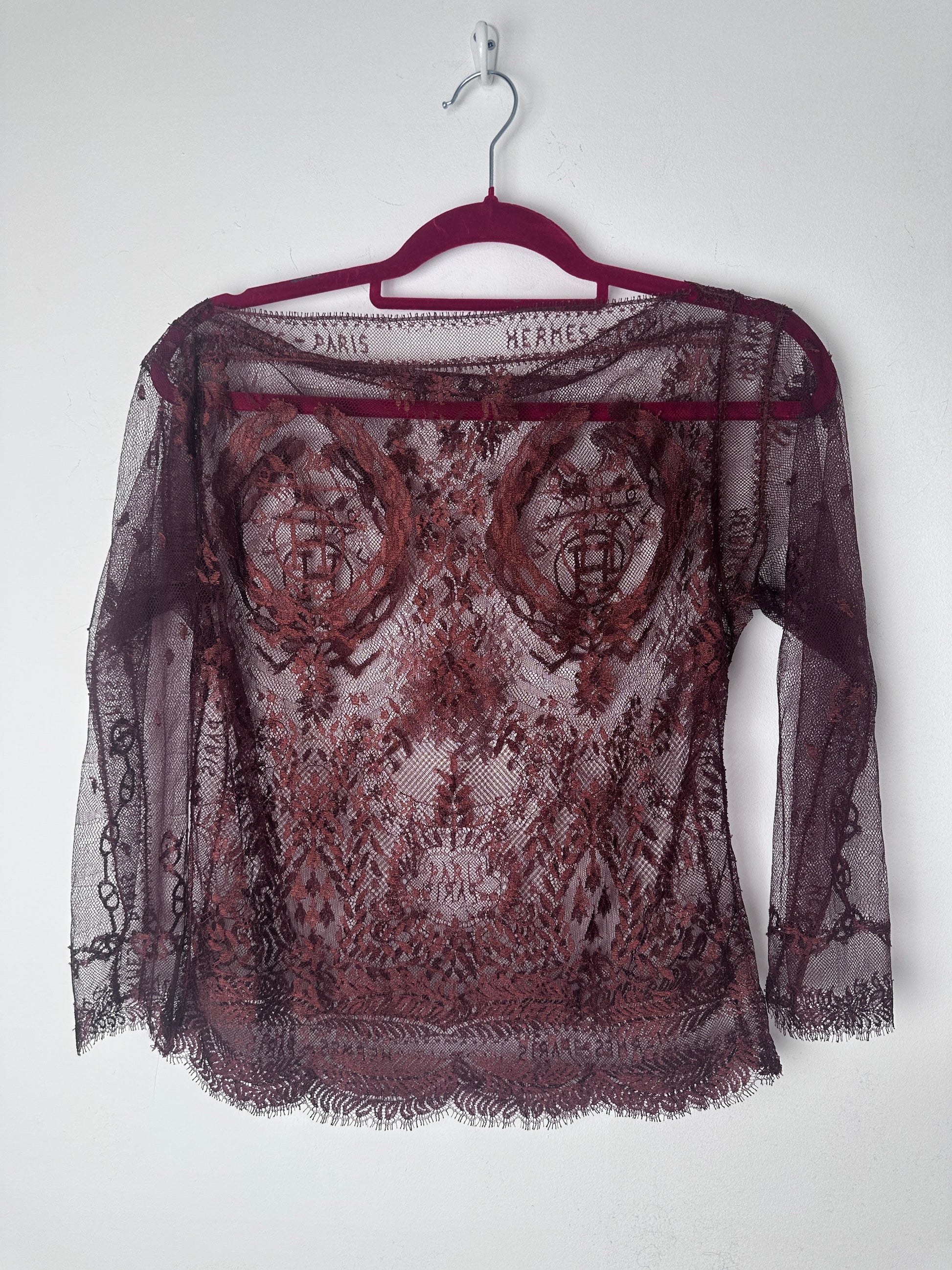 A sheer burgundy lace shawl with a floral pattern and fringed edges, draped over a matching burgundy hanger against a plain white background.