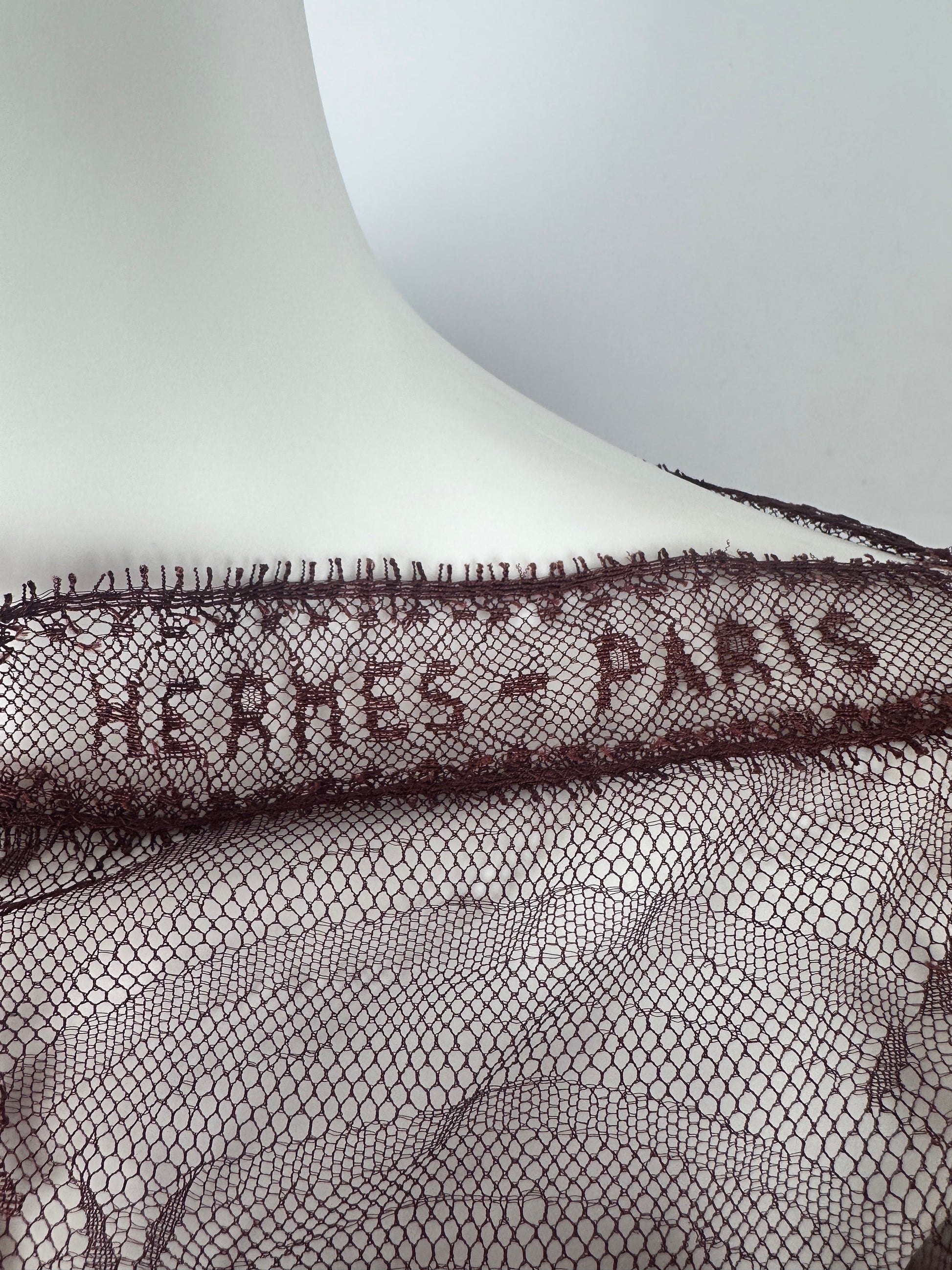 Close-up of a dark brown Hermès Paris lace trim draped over a white mannequin torso, showing the embroidered brand name and mesh pattern.