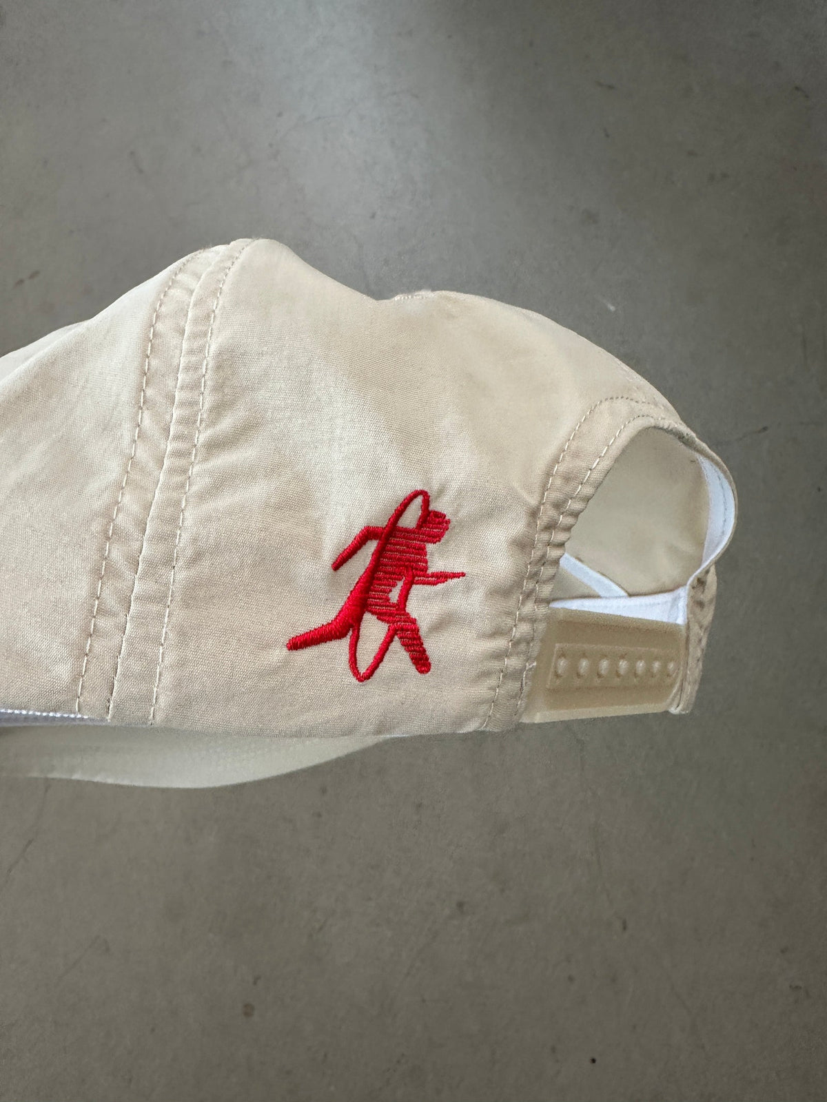 A beige baseball cap with a red embroidered running figure logo on the side, photographed against a gray concrete floor.