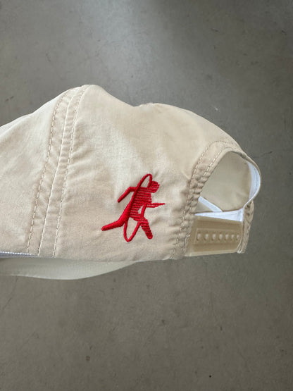 A beige baseball cap with a red embroidered running figure logo on the side, photographed against a gray concrete floor.