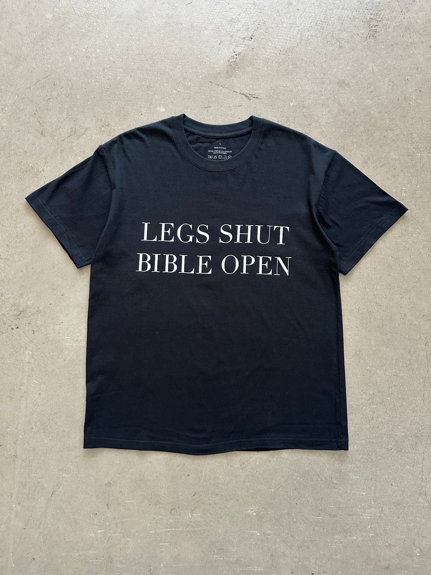 Black t-shirt with text 'LEGS SHUT BIBLE OPEN' on a concrete surface