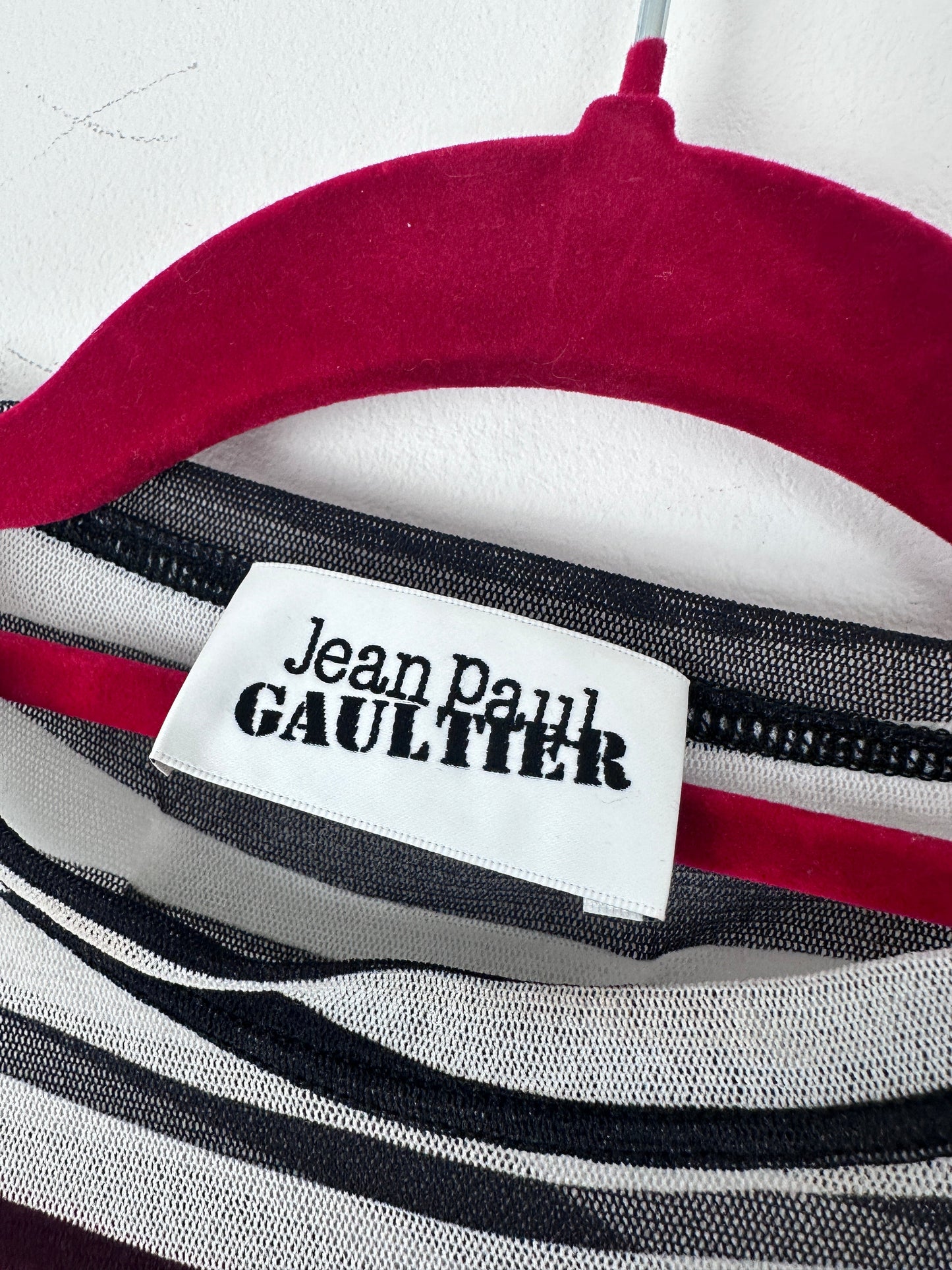 Close-up of a striped garment with a white fabric label reading 'Jean Paul Gaultier' in black text, showing red, white, black, and gray stripes.