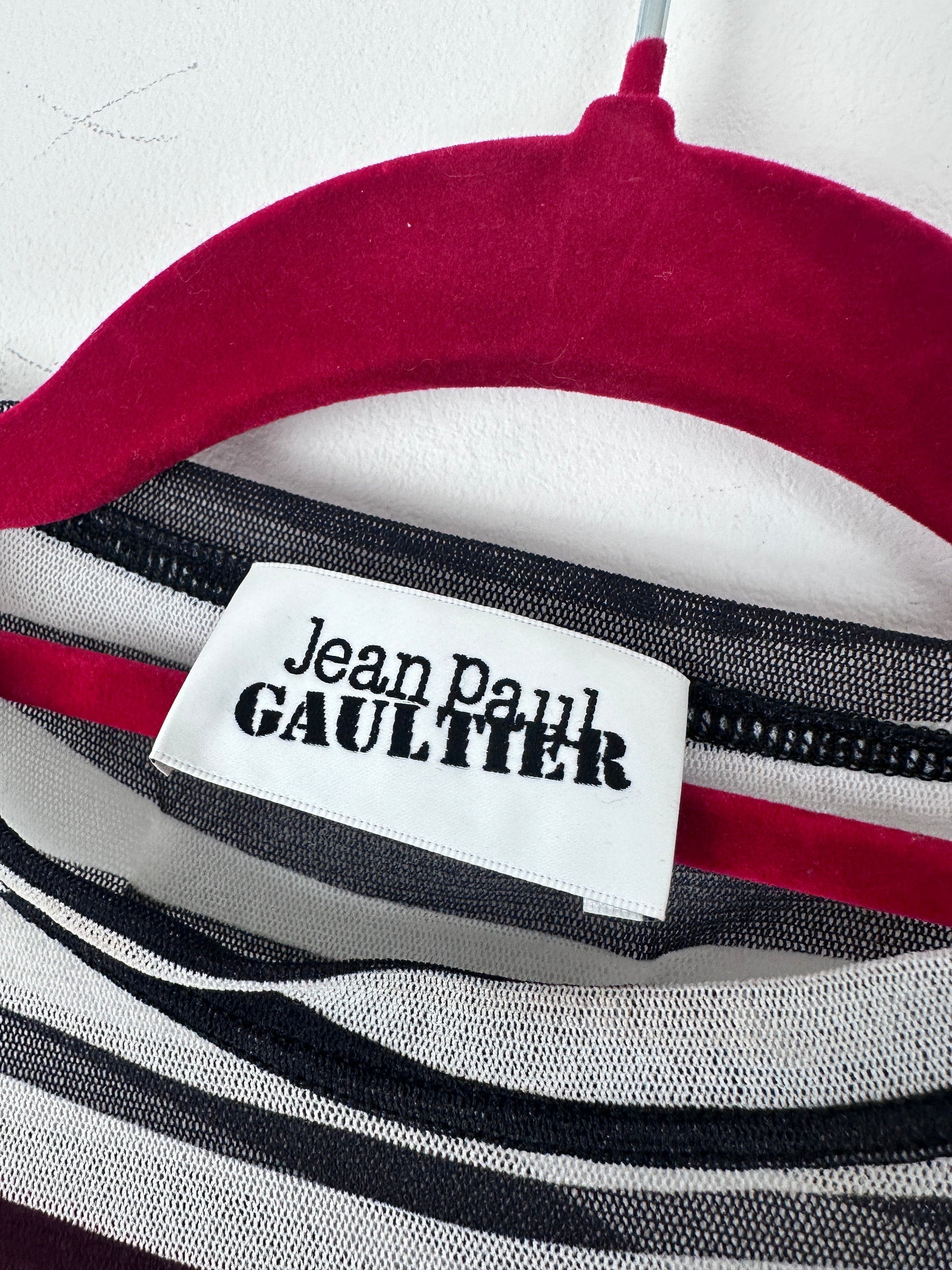 Close-up of a striped garment with a white fabric label reading 'Jean Paul Gaultier' in black text, showing red, white, black, and gray stripes.