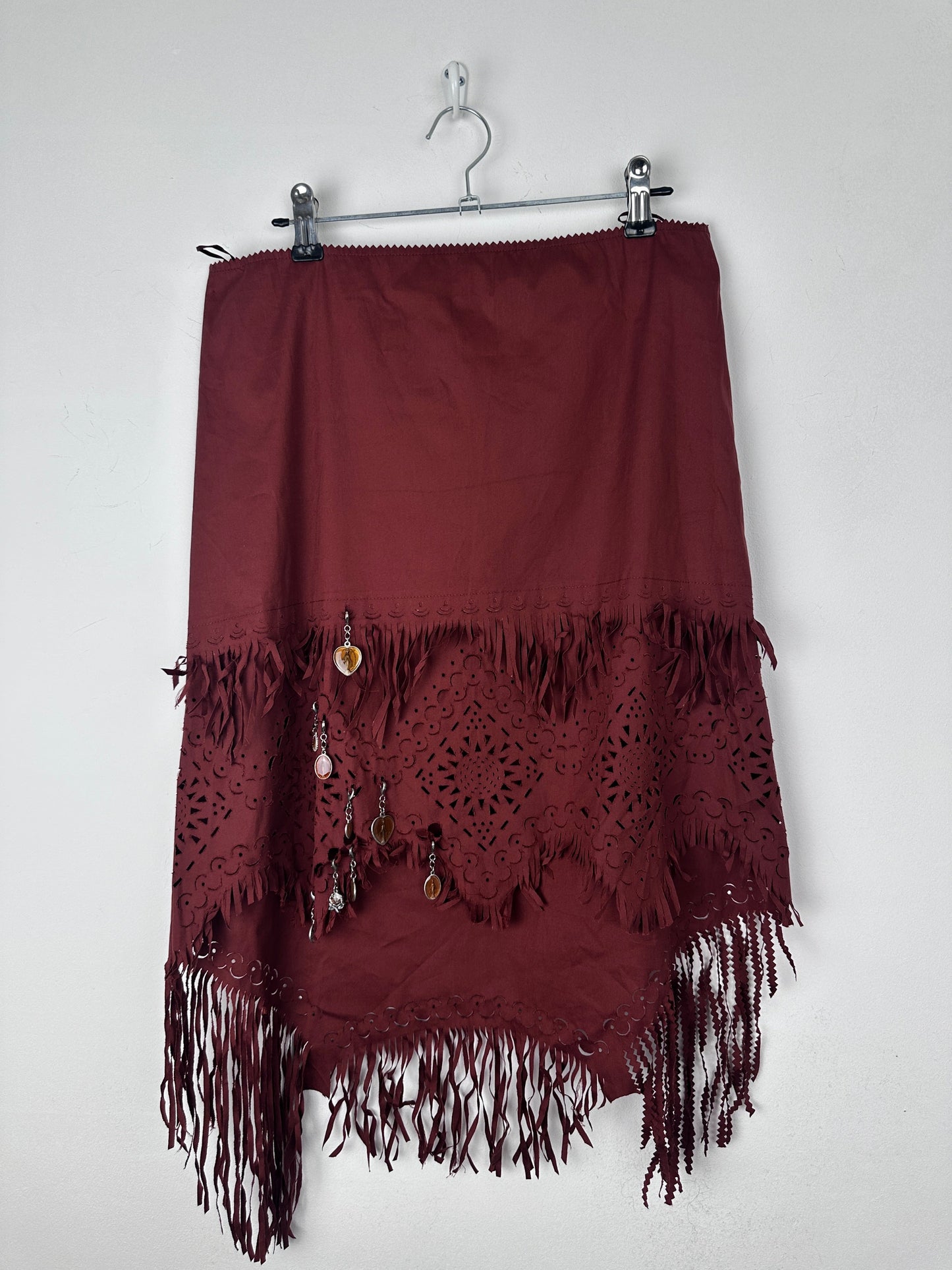 A dark burgundy skirt with intricate lace cutouts and fringe trim, hanging on a metal hanger against a plain white background.