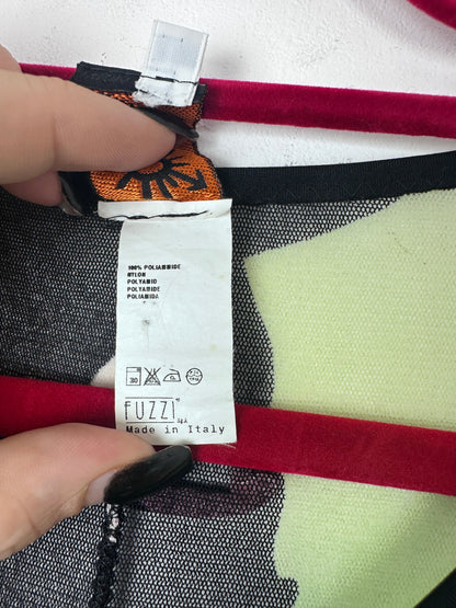 Close-up of a garment label reading FUZZI Made in Italy, held by two hands against a patterned textile background with red, black, white, and pale yellow sections.