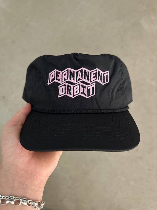 A black snapback cap with 'PERMANENT ORBIT' embroidered in pink and white on the front, held by a hand against a neutral concrete wall background.
