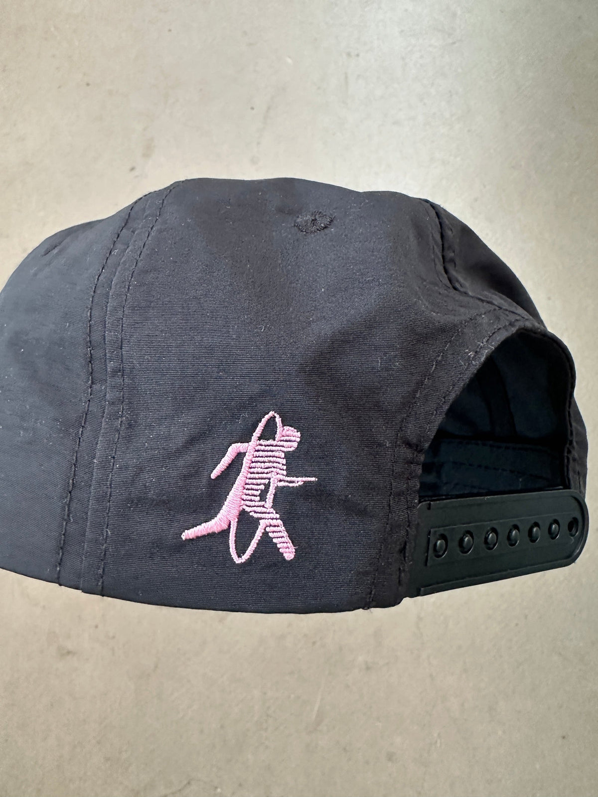 A black baseball cap with a pink embroidered graphic of a running figure holding a rifle, shown from the back with the adjustable strap visible against a neutral concrete background.