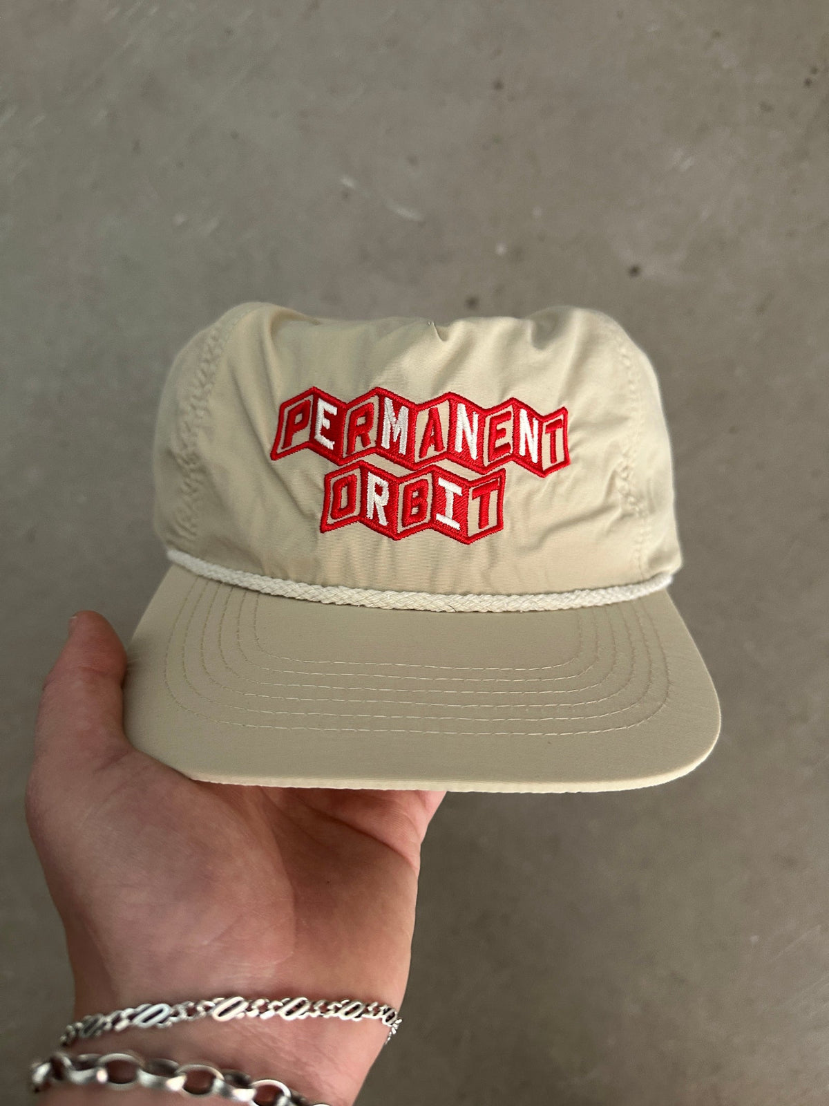 A beige Permanent Orbit baseball cap held by a hand, featuring red embroidered 3D-effect lettering on the front panel.