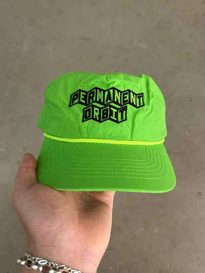 A hand holding a bright neon green baseball cap with 'PERMANENT ORBIT' embroidered in black and green on the front, set against a neutral concrete background.