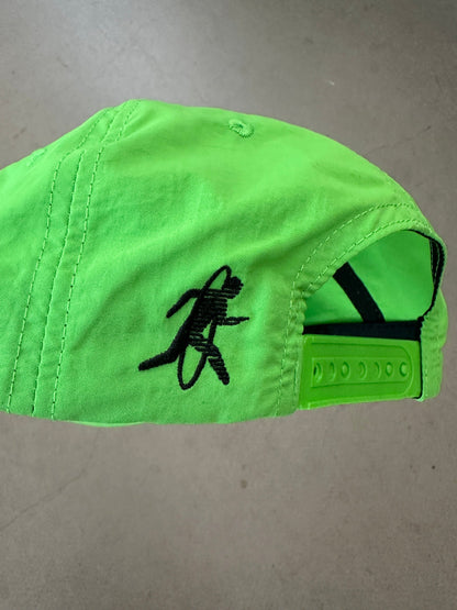 A bright neon green baseball cap with a black embroidered running figure logo on the side, photographed on a neutral gray surface.