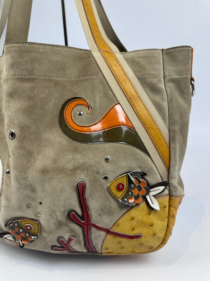 A taupe suede shoulder bag with a wide tan shoulder strap, decorated with colorful fish-shaped enamel pins and a yellow ostrich-quill patch on the side.